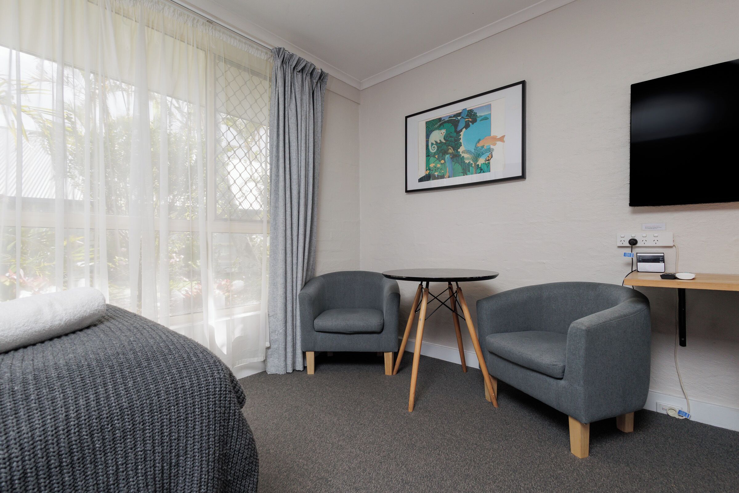 Quarto de hotel com uma cama, duas poltronas, uma mesinha, televisão e um quadro na parede.