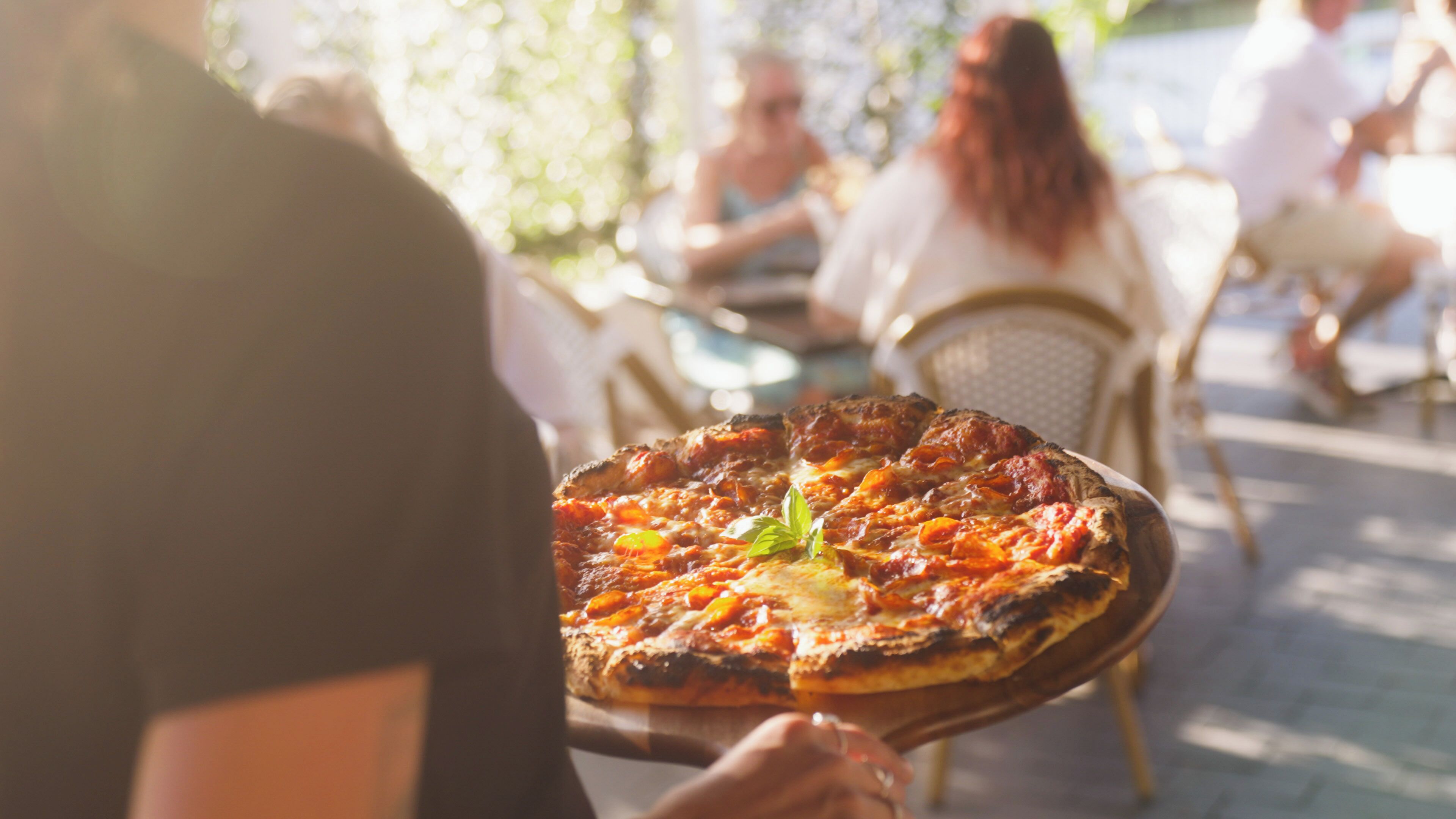 Una persona sosteniendo una pizza adornada con una hoja de albahaca, sentada en una mesa al aire libre con otros comensales de fondo.