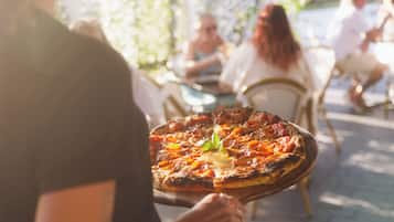 Una persona sosteniendo una pizza adornada con una hoja de albahaca, sentada en una mesa al aire libre con otros comensales de fondo.