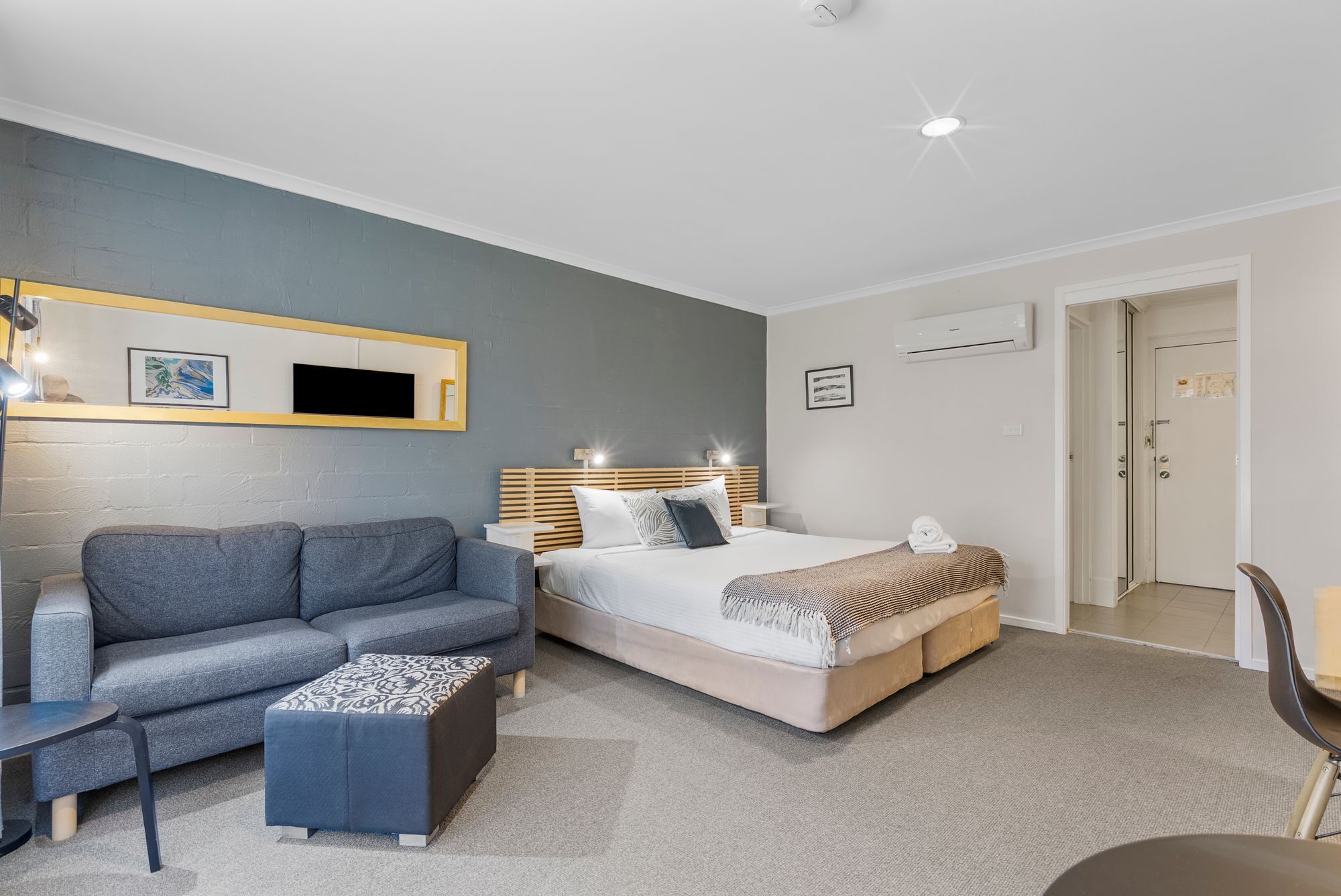 Une chambre d’hôtel moderne avec un canapé gris, un lit doté d’une tête de lit en bois et une petite table.