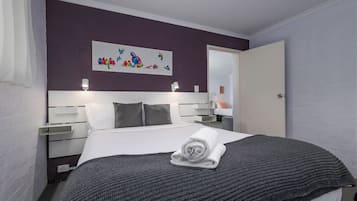 A neatly made bed with a rolled towel, a headboard with wall art, bedside lamps, and a door to the right.