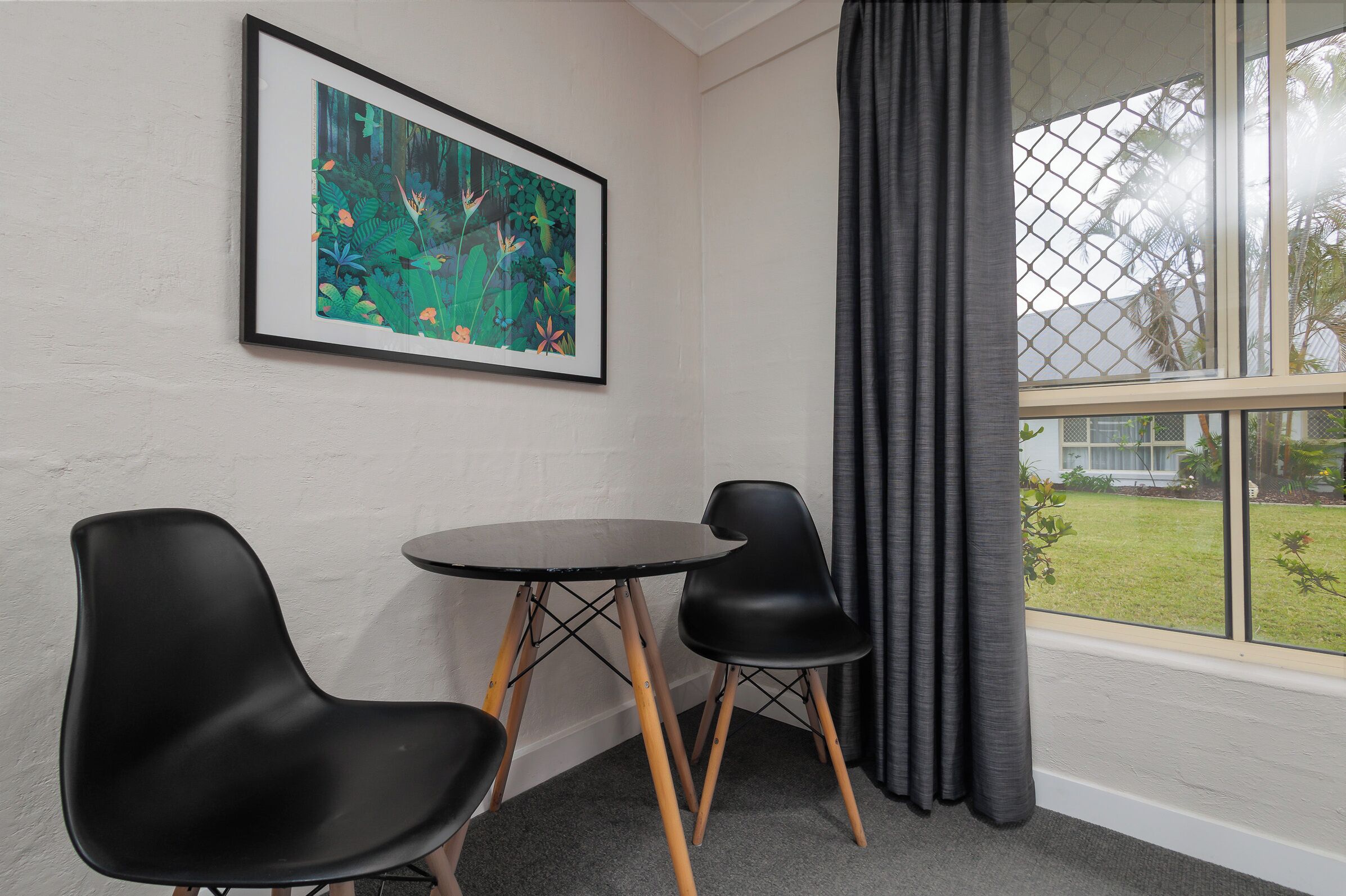A room with a framed painting, two black chairs, a small round table, and a window with a view of greenery.