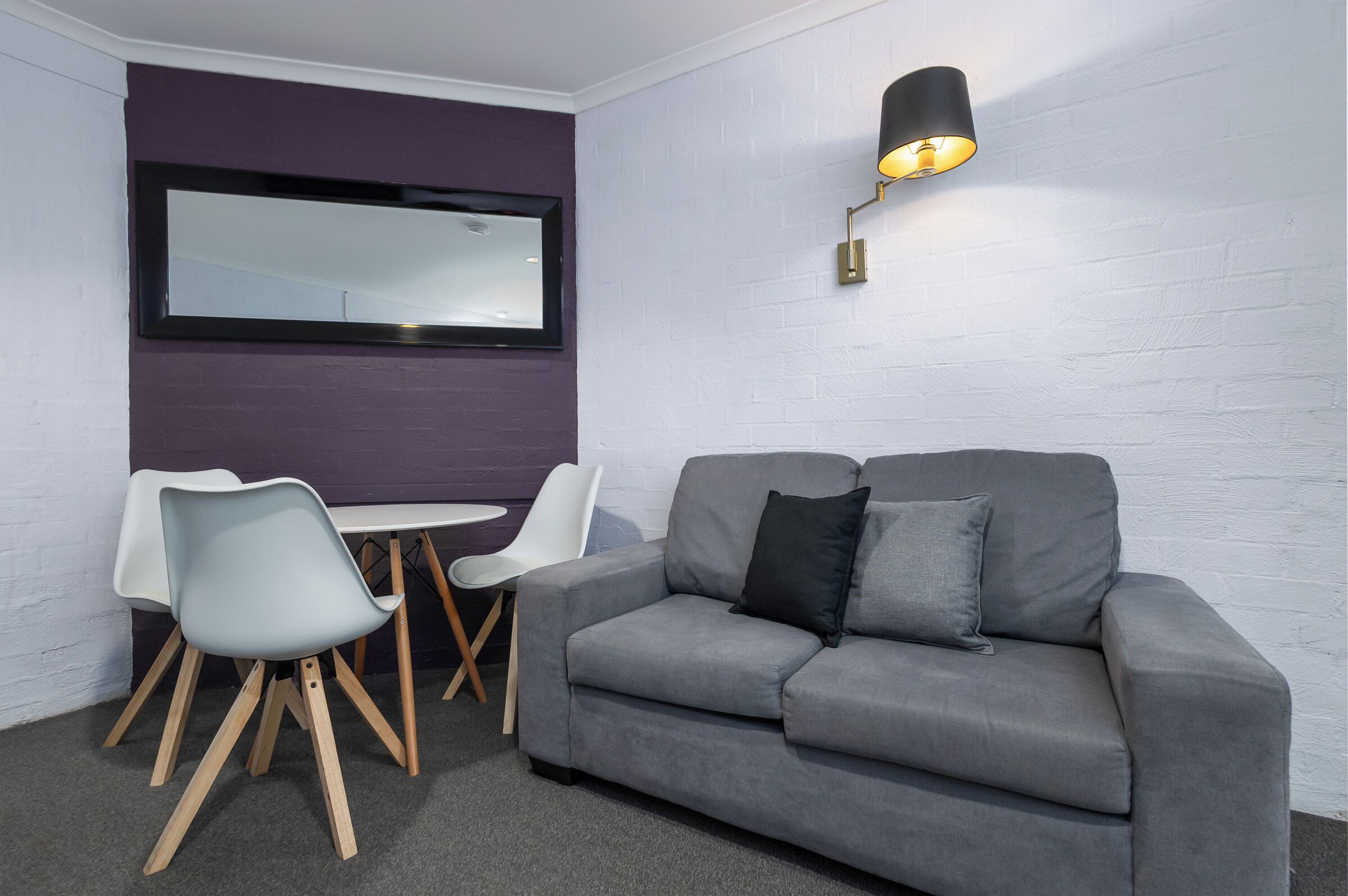 Een moderne kamer met een grijze bank, twee witte stoelen, een klein tafeltje en een wandlamp.