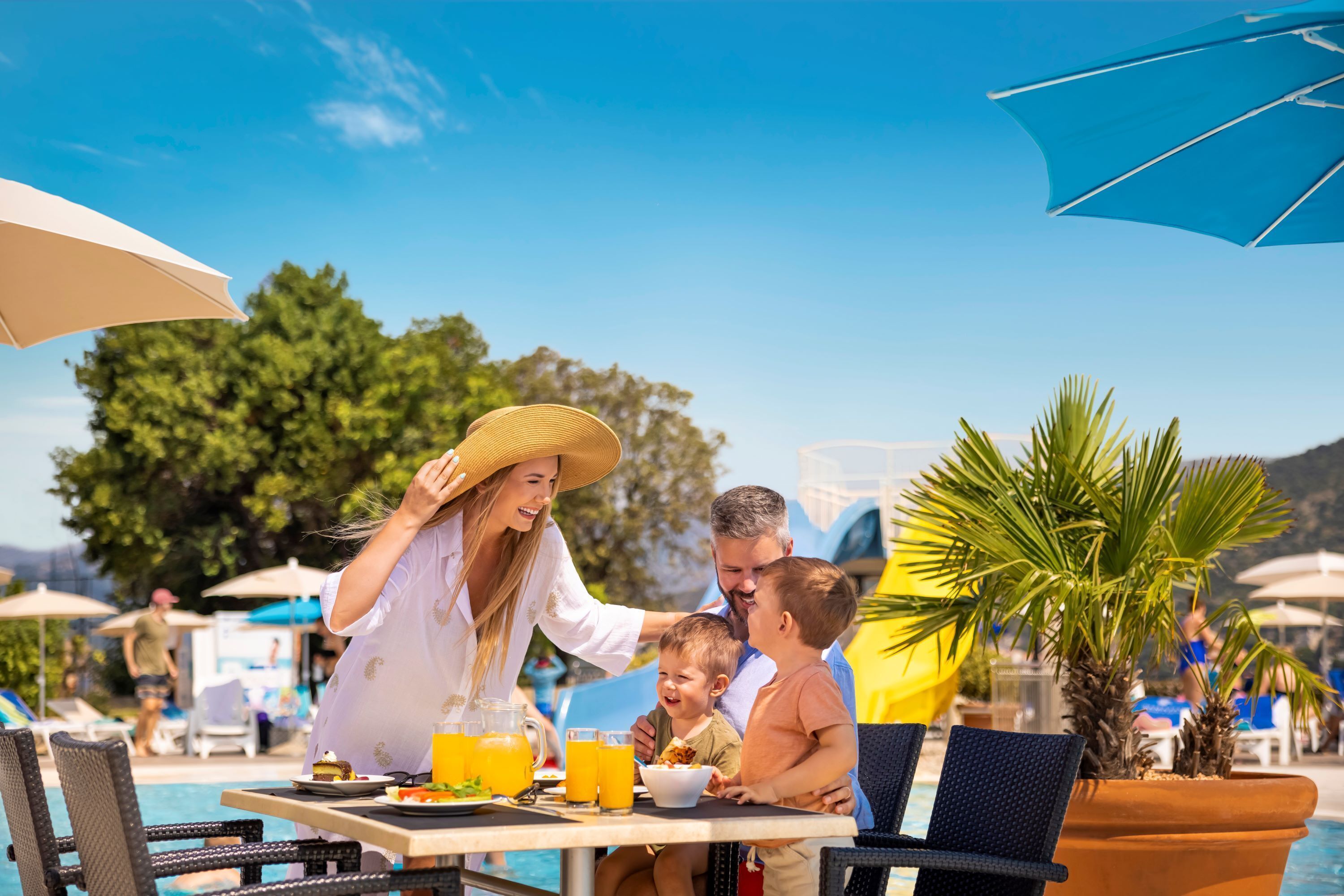 Una familia cenando en una mesa junto a la piscina, con vistas a la piscina y palmeras.