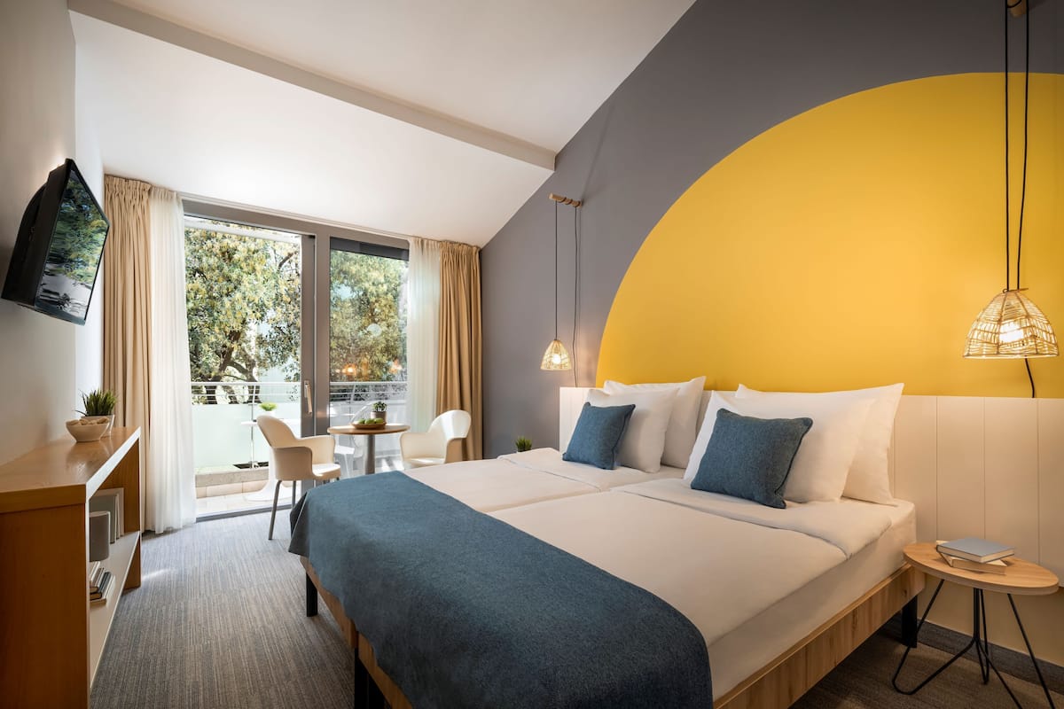 A modern hotel room with a large bed, a desk, a chair, and a balcony with a view of trees.