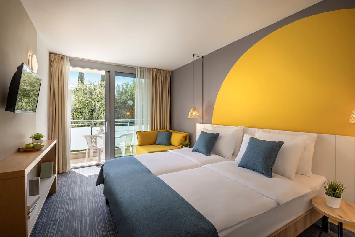 A hotel room with a large bed, a desk, a chair, and a balcony with a view of trees.