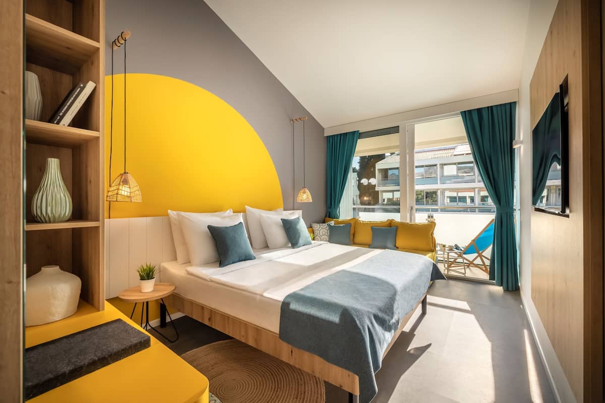 A modern hotel room with a bed, bedside tables, a small seating area, a bookshelf, and a large window with curtains.