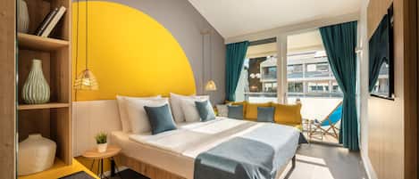 Una habitación de hotel moderna con cama, mesitas de noche, un pequeño rincón de descanso, una estantería y un amplio ventanal con cortinas.