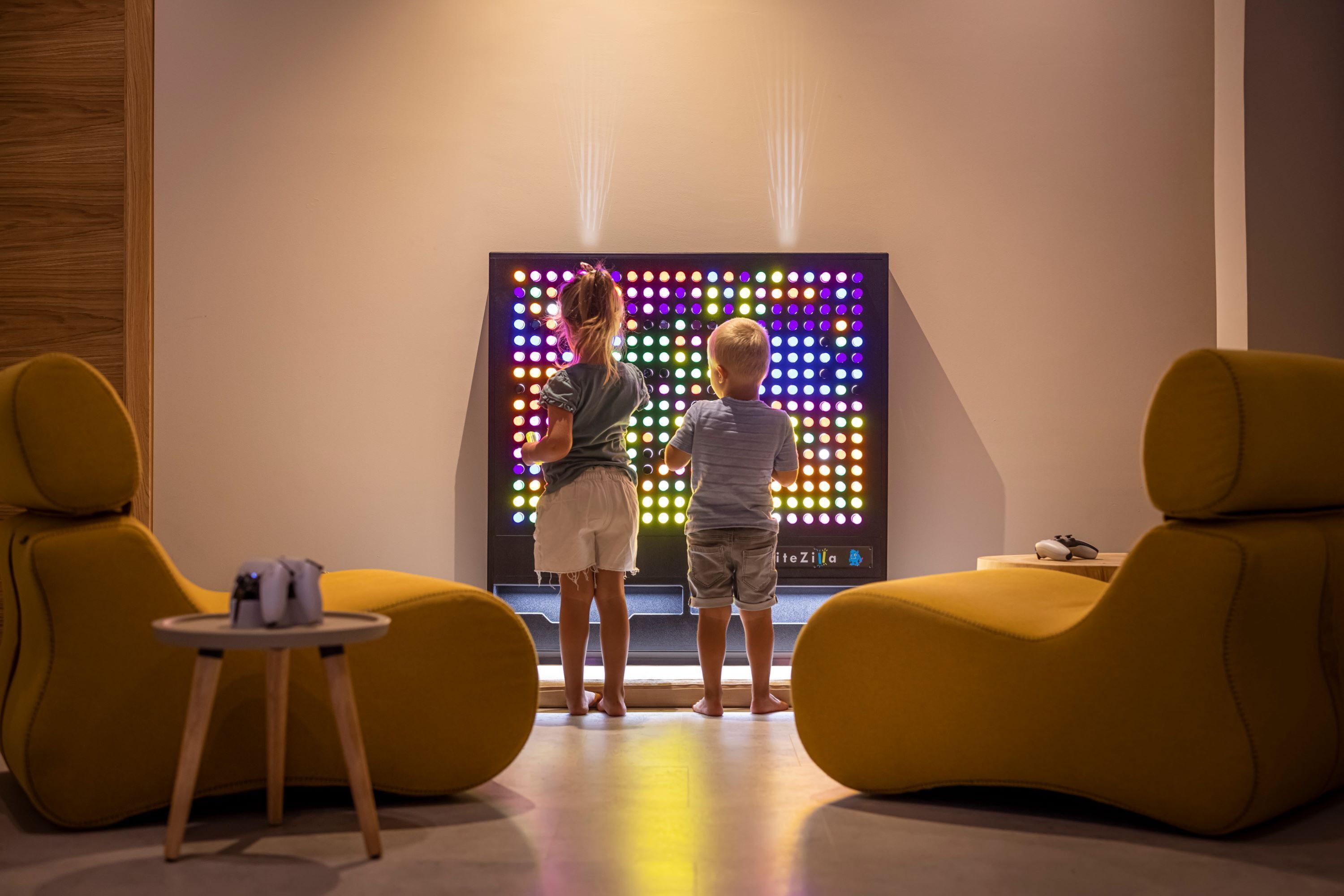 Two children standing in front of a colorful LED display in a modern room with yellow chairs and a small table.