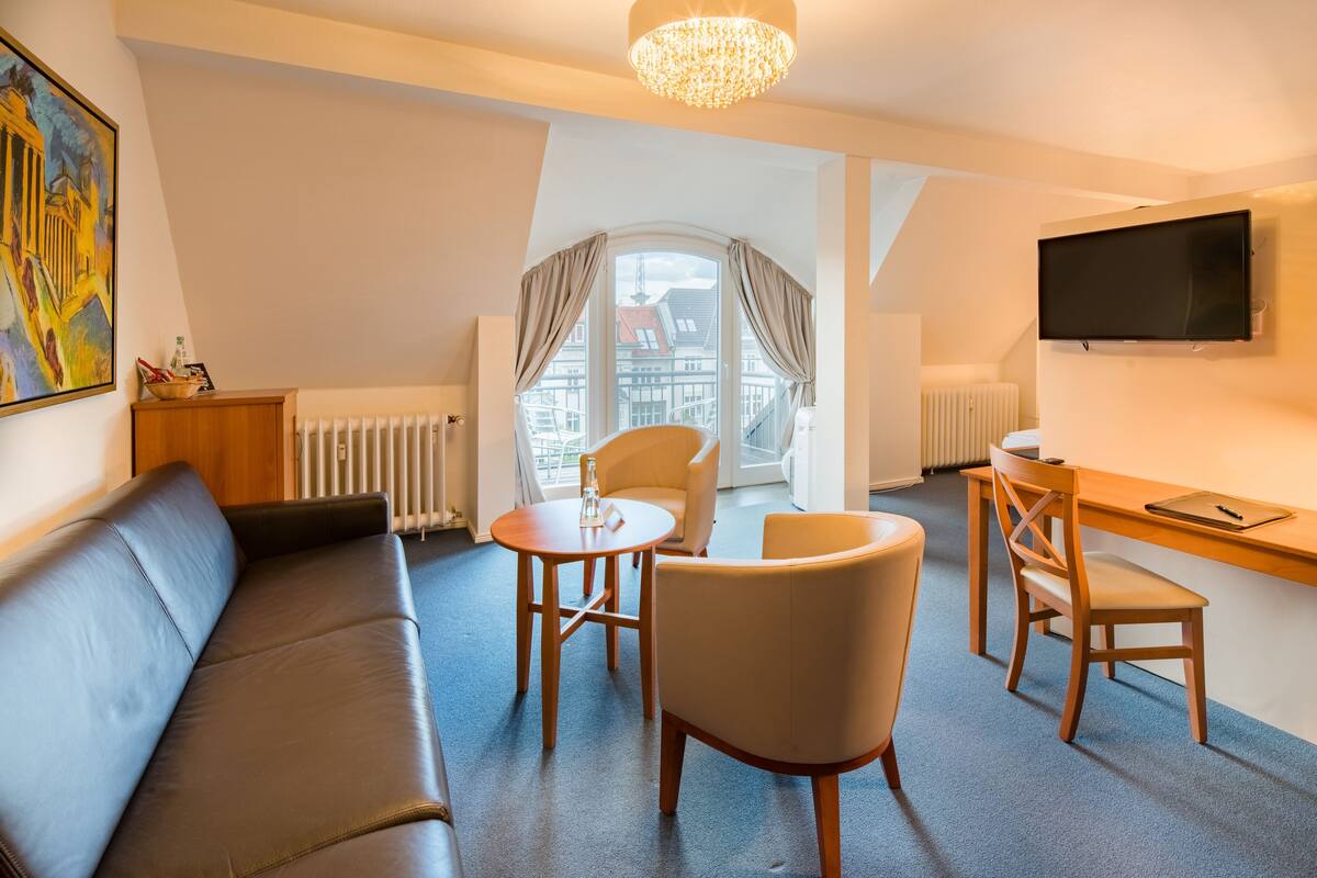 Una habitación de hotel moderna con sofá, dos sillones, una mesita, televisión y ventana con cortinas.