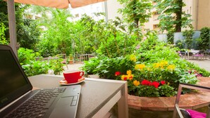 A laptop on a table with a red cup beside it, surrounded by a garden with various plants and flowers. - Hotel Brandies Berlin (Berlin)