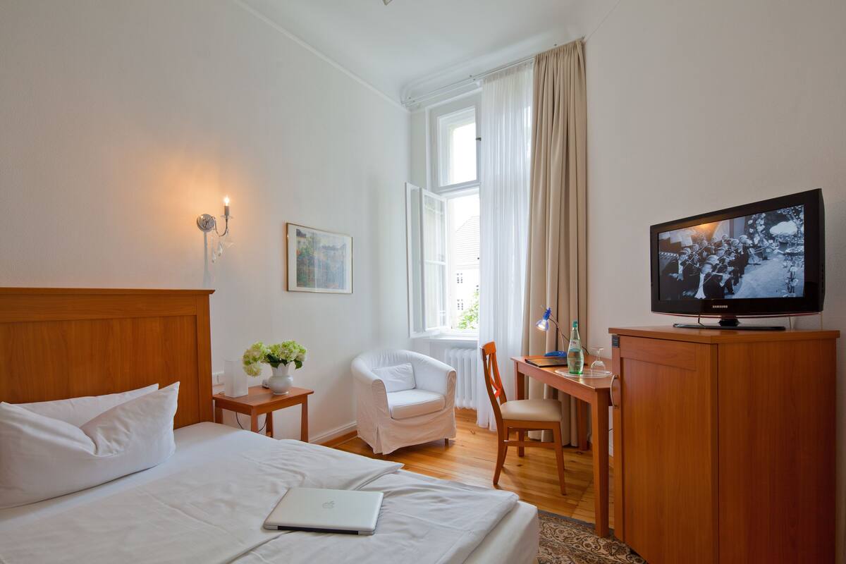 Habitación de hotel con cama, escritorio con una computadora portátil, silla, televisor y ventana con cortinas.