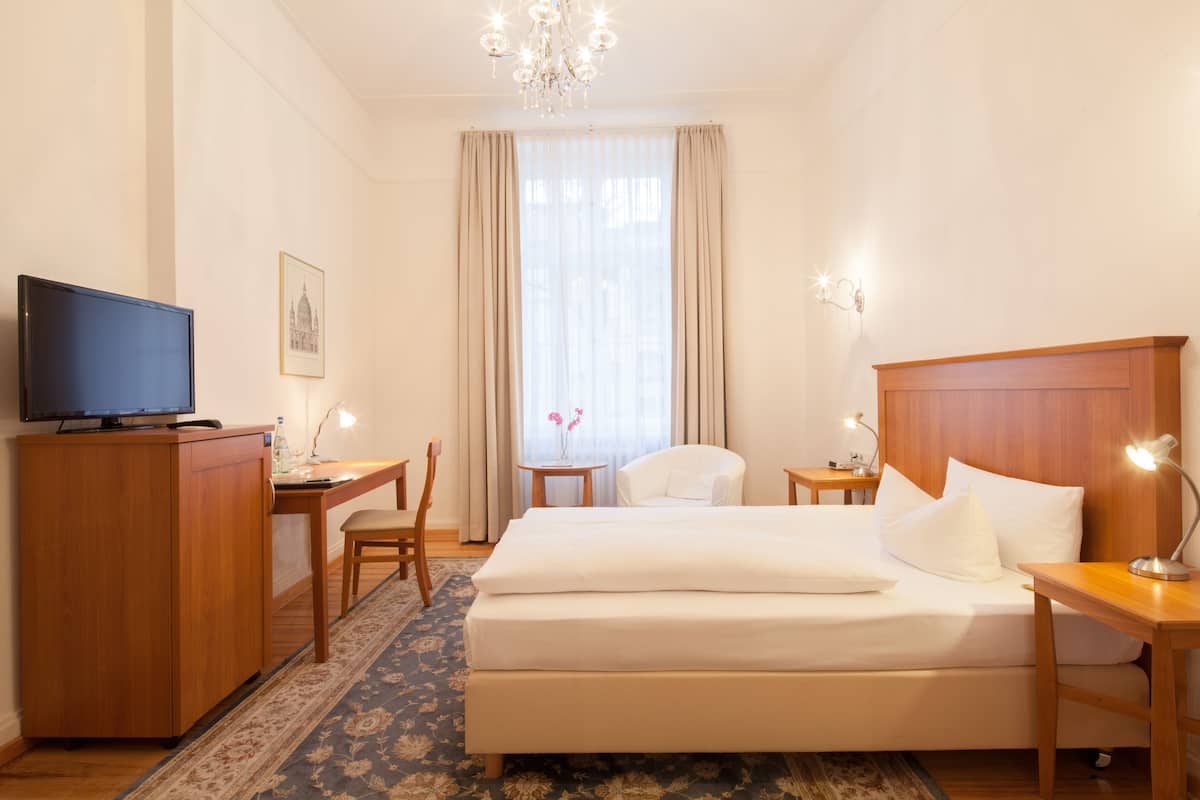 Una habitación de hotel con cama, escritorio, televisor, silla, una mesita y una araña.