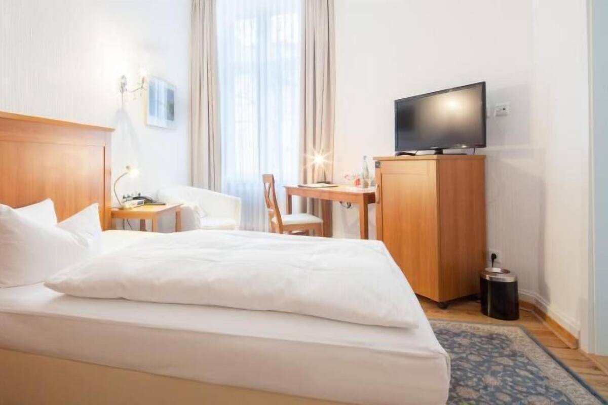 Habitación de hotel con cama, escritorio, silla, televisión y cesto de basura.
