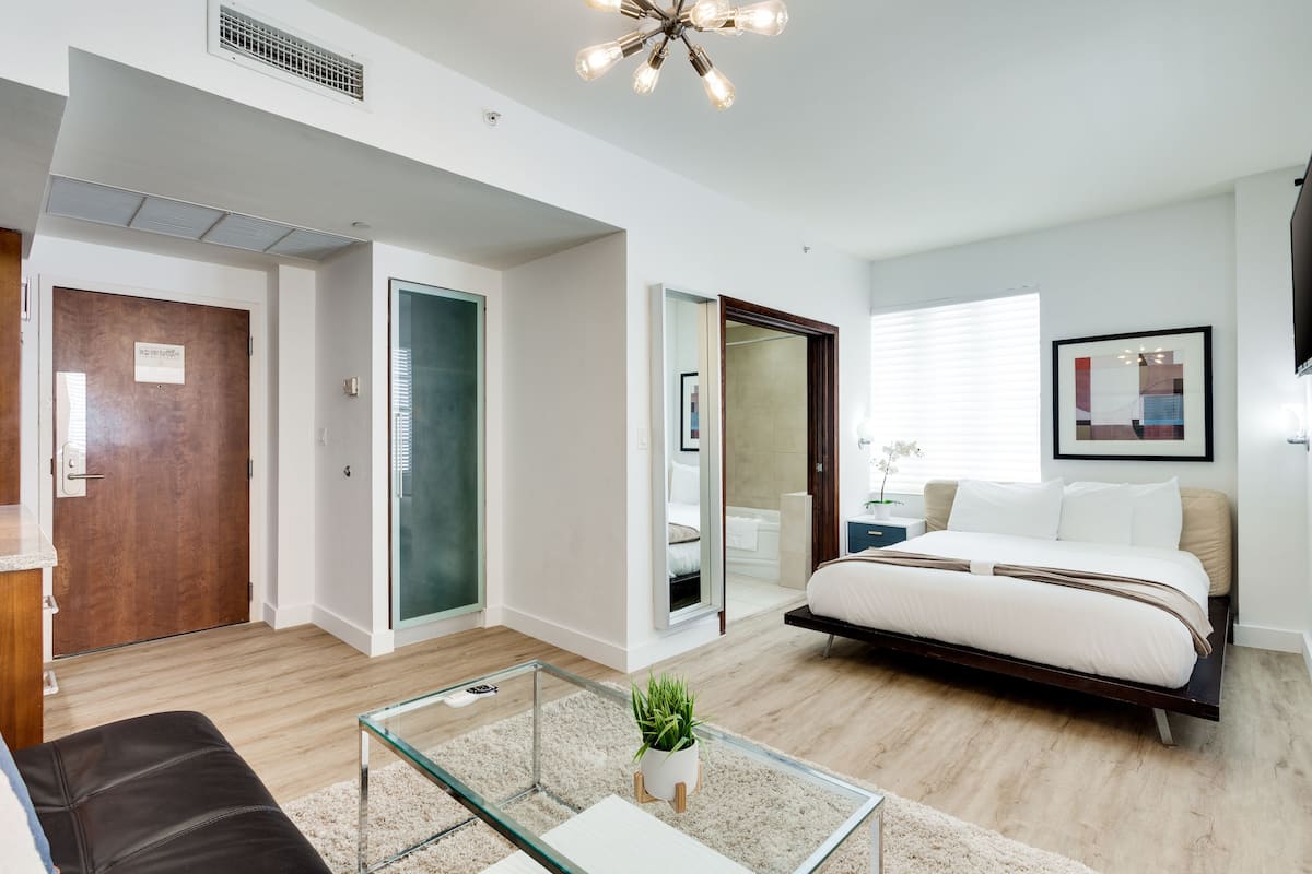 A modern hotel room with a bed, a glass coffee table, a sofa, and a bathroom visible through an open door.