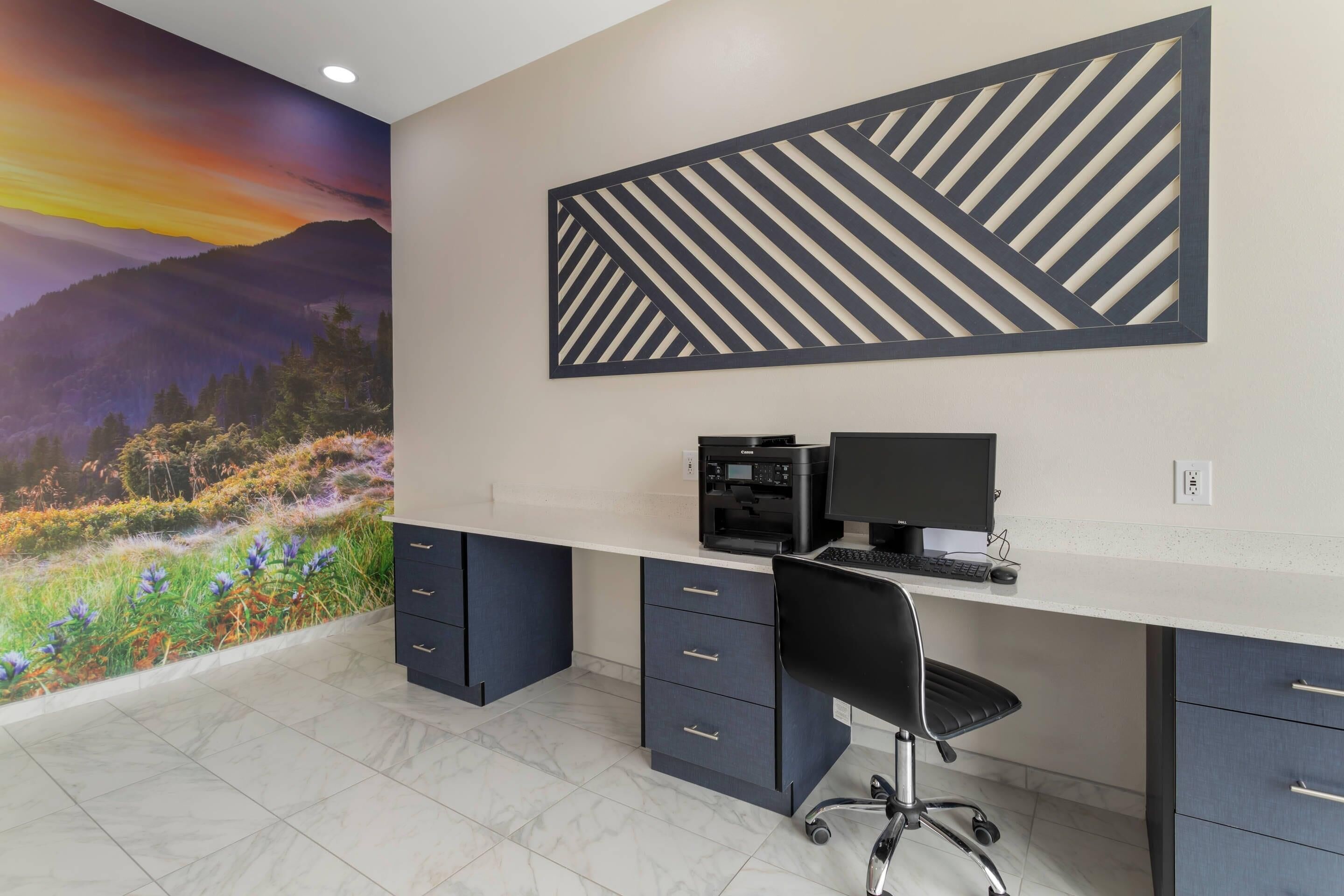 A modern office space with a desktop computer, a chair, and a decorative wall with a nature scene and geometric patterns.