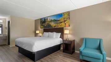 Una habitación de hotel con una cama grande, una mesita de noche con lámpara, un sillón azul turquesa y un cuadro de un paisaje en la pared.