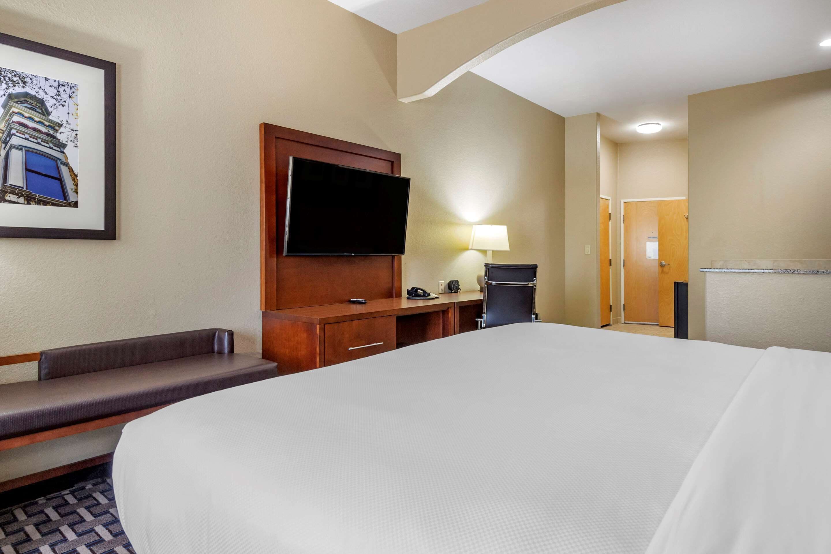 A hotel room with a large bed, a flat-screen TV mounted on the wall, a desk with a lamp, and a bench.