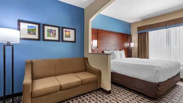 Una habitación de hotel con un sofá marrón, una cama con ropa blanca, una mesita de noche con una lámpara y cuadros en la pared.