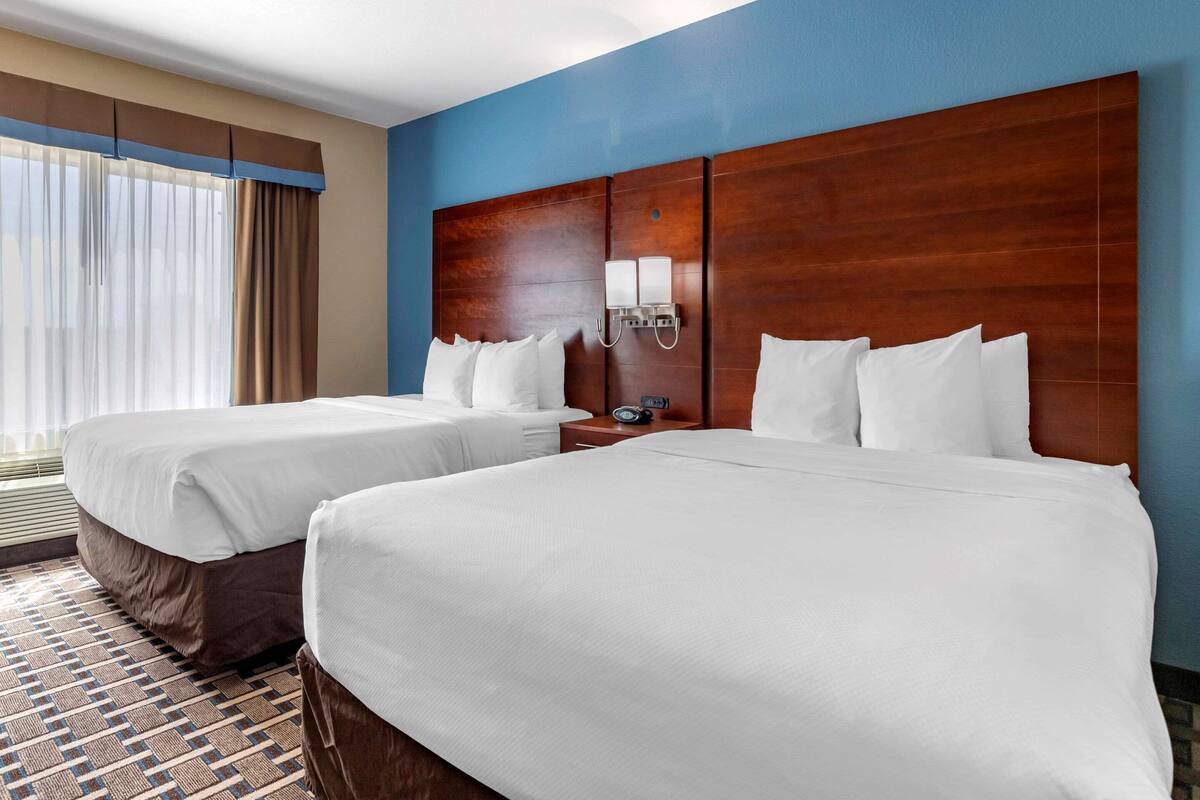 A hotel room with two beds, a wooden headboard, a nightstand with a clock, and a window with curtains.