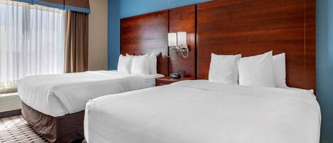 A hotel room with two beds, a wooden headboard, a nightstand with a clock, and a window with curtains.