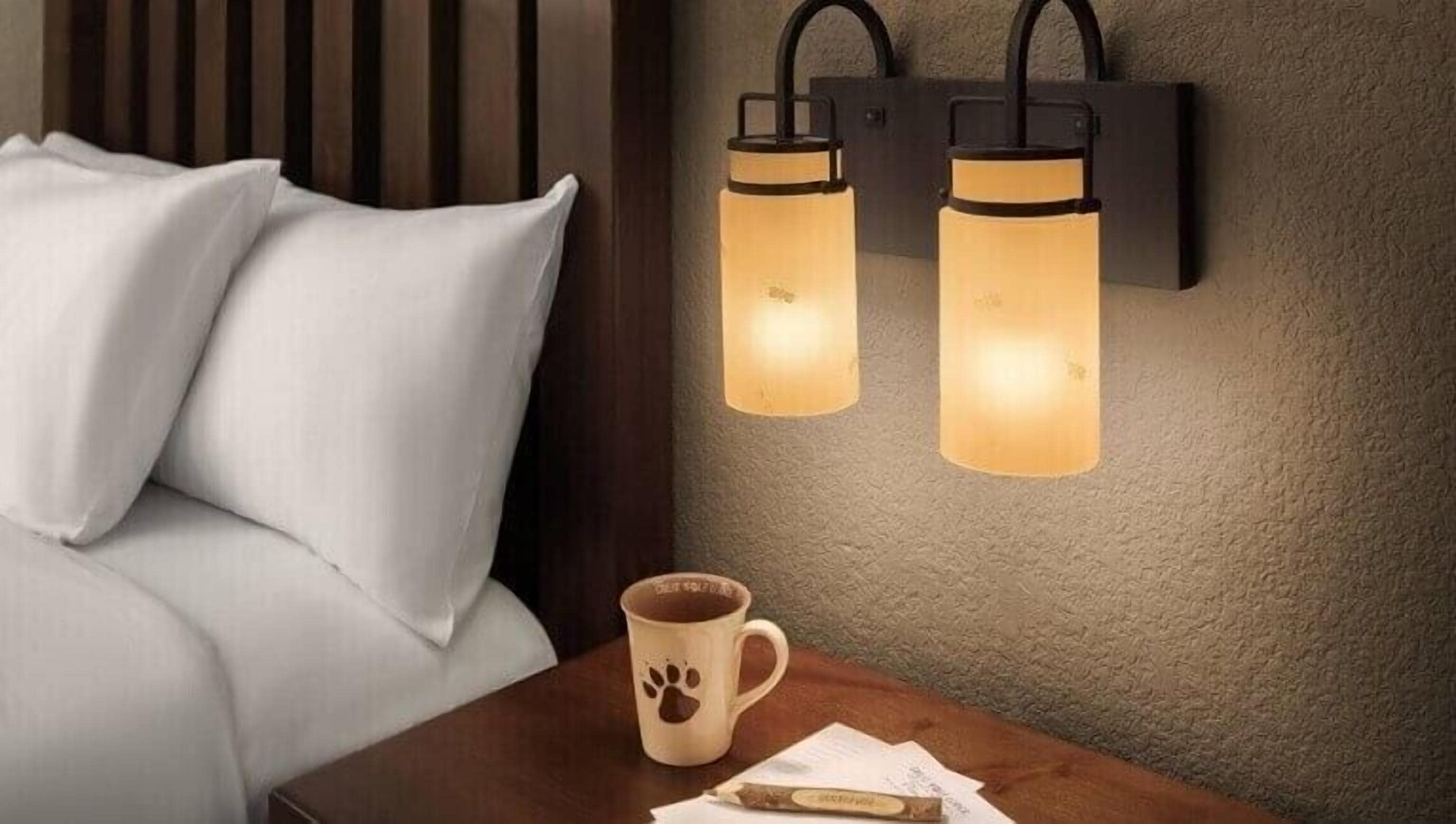 A bed with white bedding, a wooden headboard, a bedside table with a mug, and two wall-mounted lights.