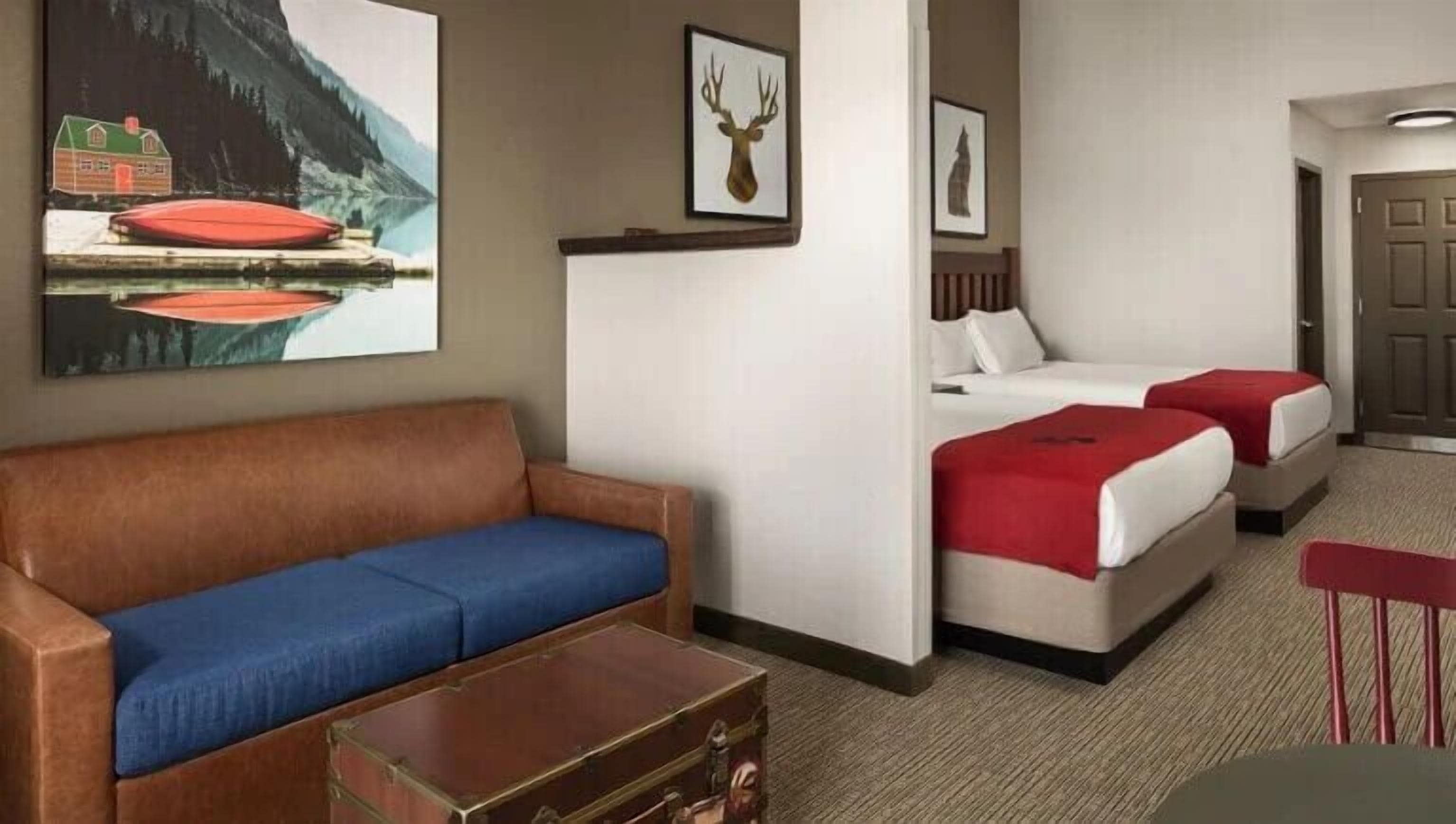 A hotel room with a brown leather sofa, two beds with red bedding, a wooden headboard, and a painting of a mountain landscape.