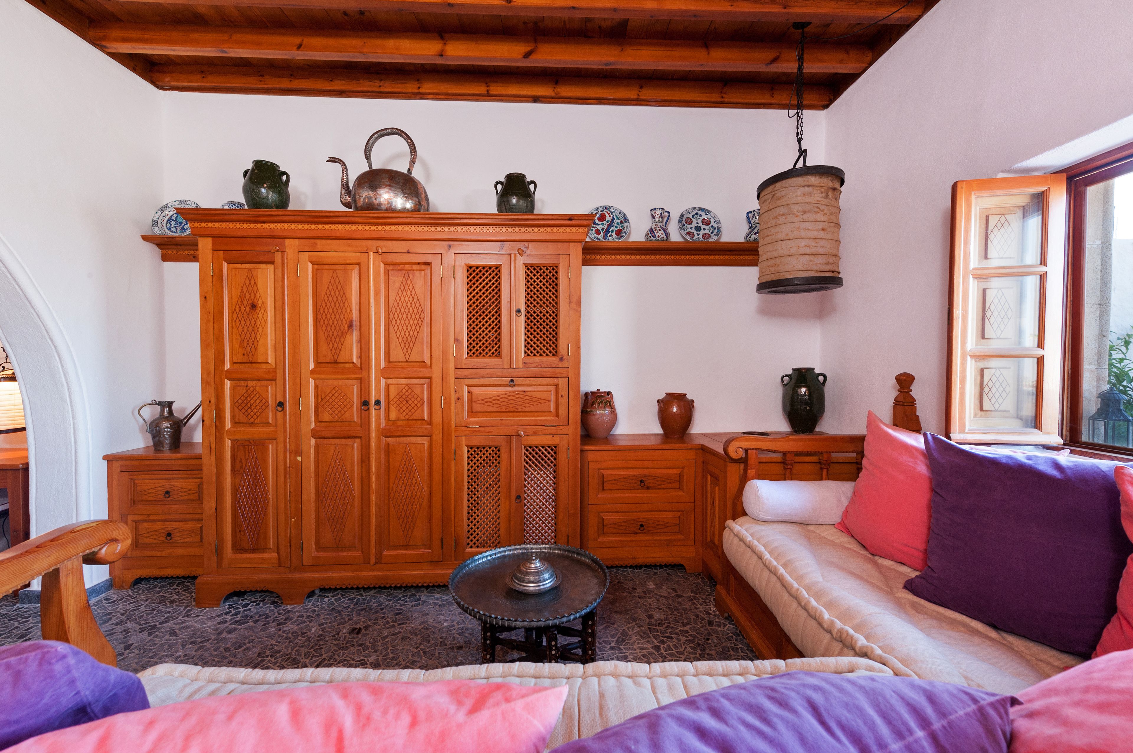 A cozy room with a wooden cabinet, a small round table, a sofa with colorful cushions, and a hanging lantern.