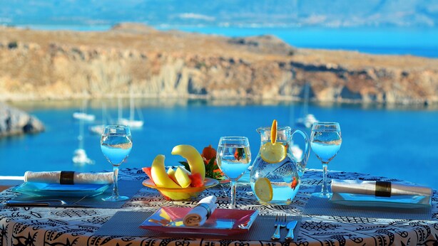 Une table dressée avec un verre de cocktail bleu, un saladier de fruits et une carafe d’eau, offrant une vue panoramique sur la côte.