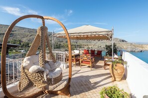 Una silla mecedora de mimbre con cojines, una pérgola de madera, mobiliario exterior y vistas al mar.
