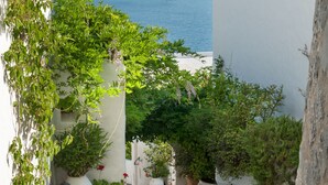 Une cour étroite agrémentée de plantes en pot, dotée d’un sol carrelé et offrant une vue sur la mer et les montagnes en arrière-plan.