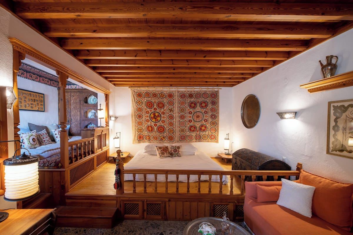 A cozy room with a wooden ceiling, a bed with a patterned headboard, a wooden bench, a stone floor, and a small table with a lamp.