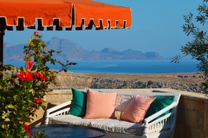 Vista al mar con un sofá blanco de mimbre bajo un paraguas naranja, cojines verdes y rosados, y una vista despejada del mar y montañas distantes.