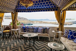 Eine Terrasse mit gemustertem Dach, einer Couch mit gemusterten Kissen, einem runden Tisch und Blick auf einen See und Berge.