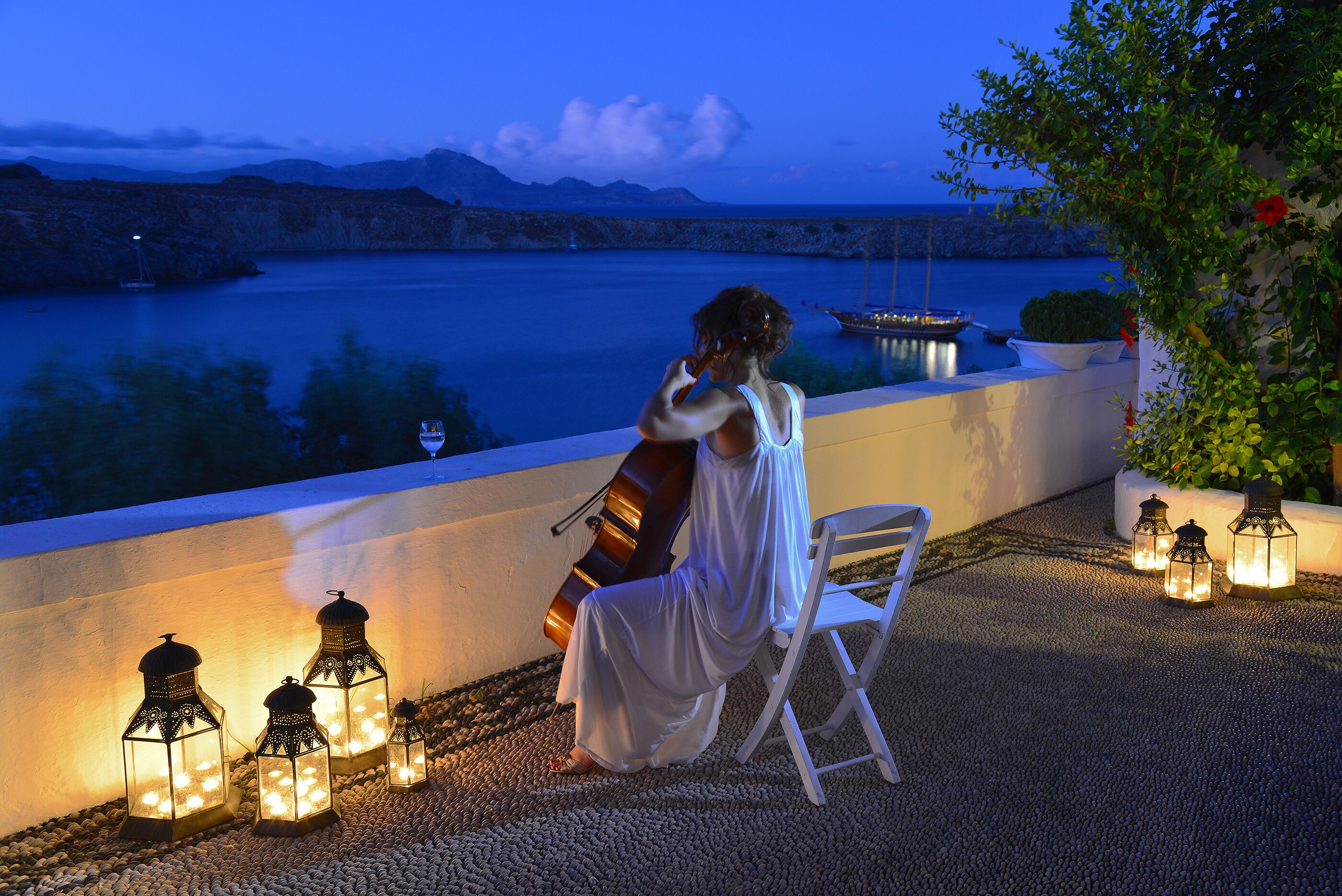 A woman in white sitting on a chair by a waterfront, overlooking a calm sea with boats and a mountainous backdrop.