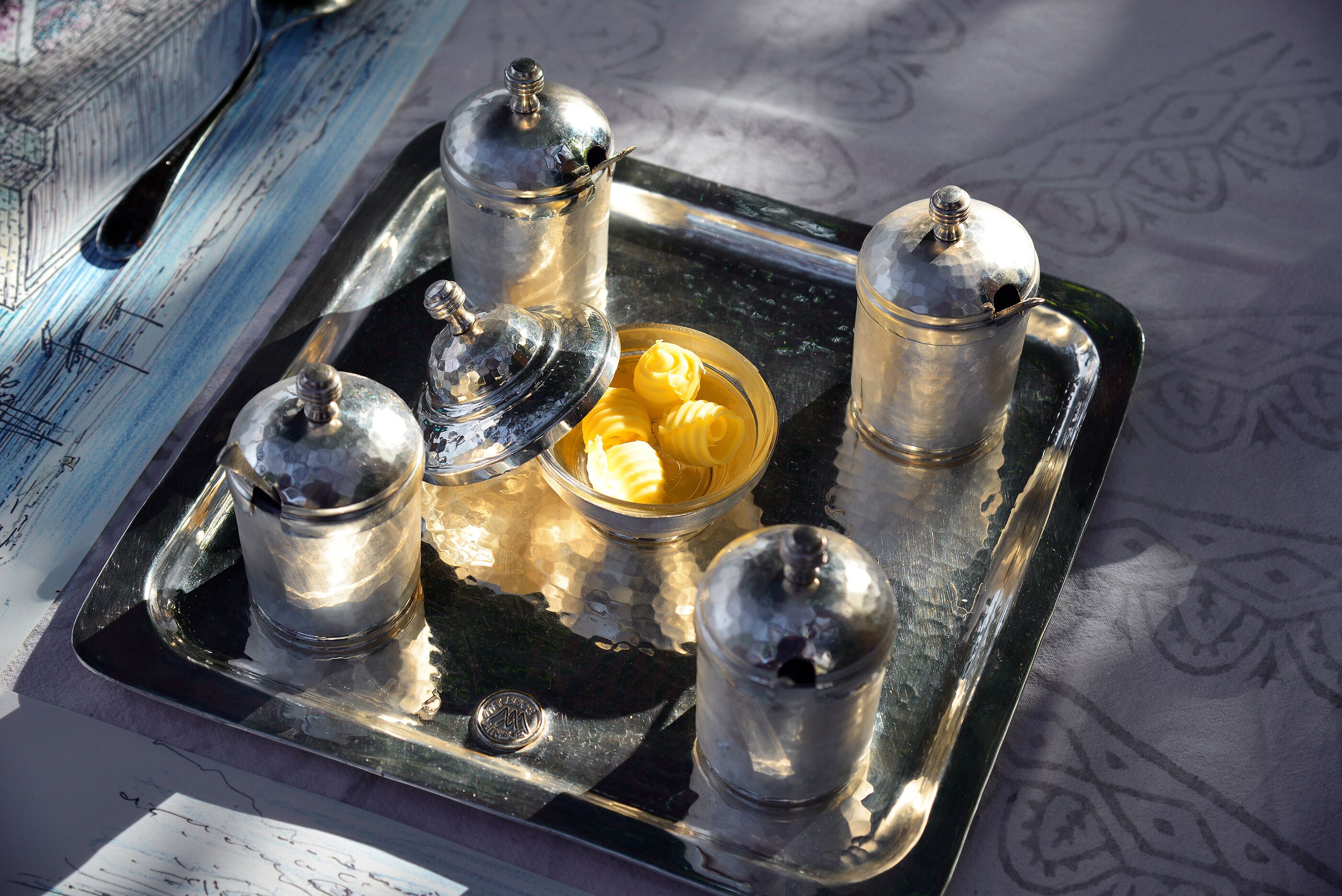 A tray with four silver containers and a bowl of yellow items, possibly food, on a table with a patterned tablecloth.