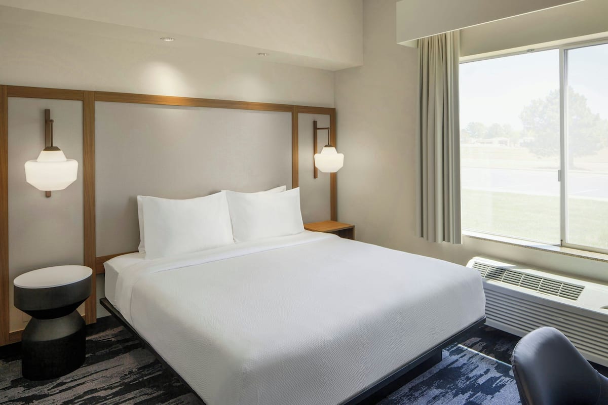 Habitación de hotel con una cama grande, dos mesitas de noche, una silla, un ventanal con vistas y una lámpara en el techo.