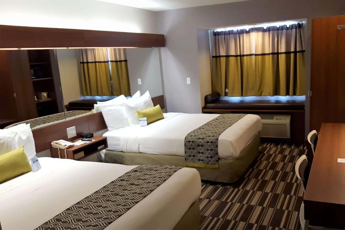 A hotel room with two beds, a desk, a chair, a mirror, and a window with curtains.