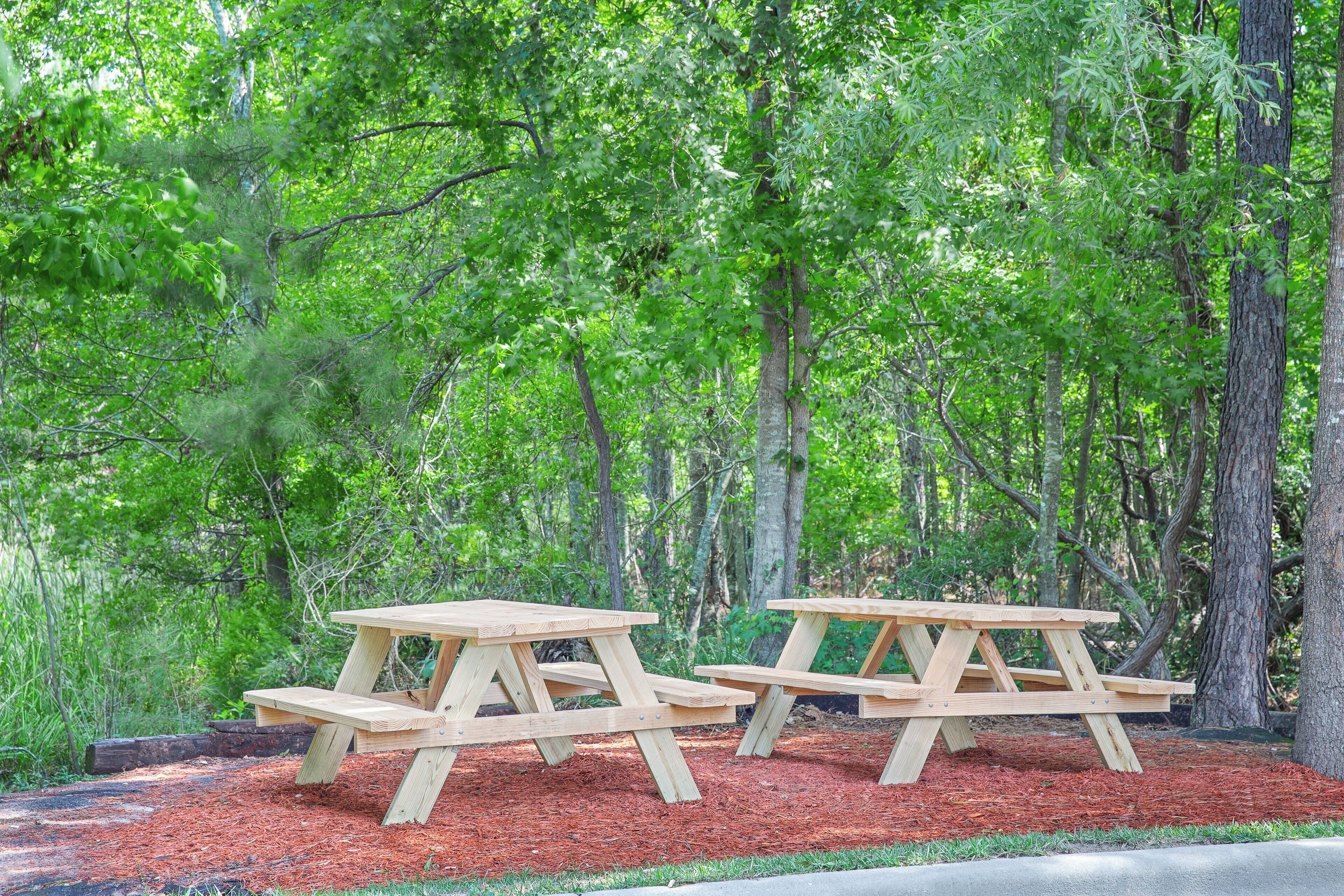Two wooden picnic tables in a forested area with mulch ground cover.