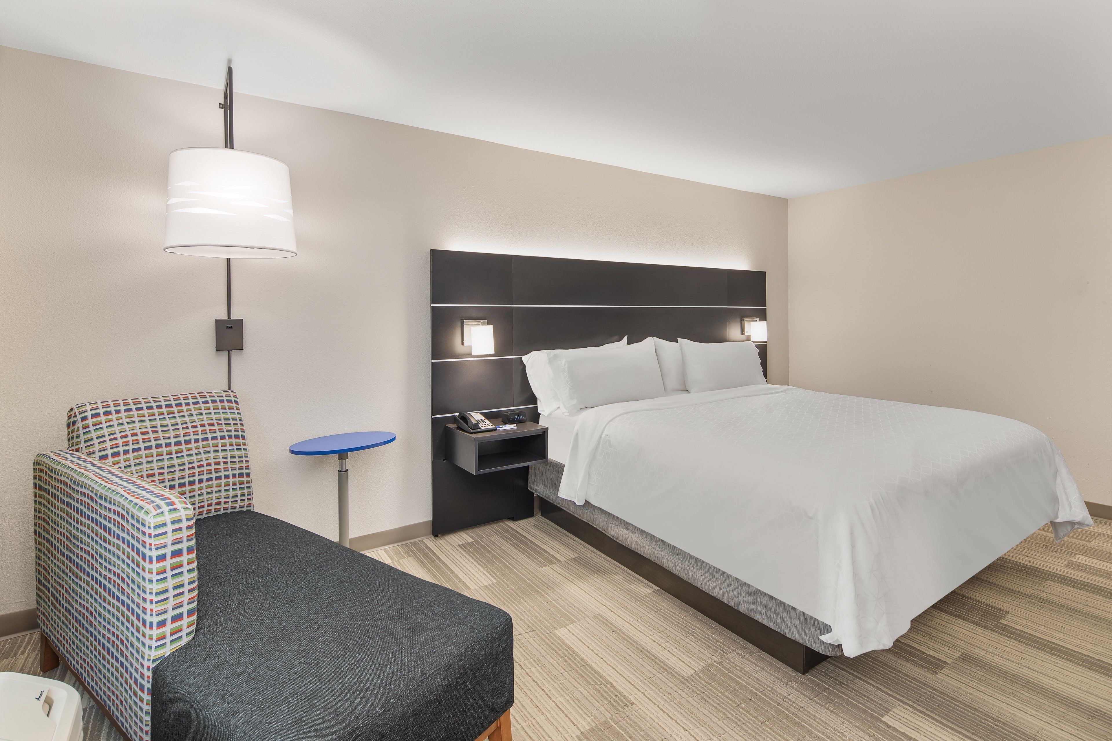 A hotel room with a bed, a chair, a small table, and a wall-mounted lamp.