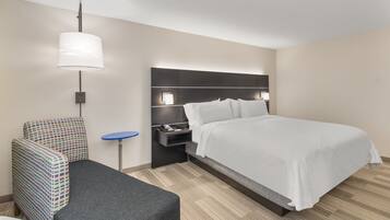 A hotel room with a bed, a chair, a small table, and a wall-mounted lamp.