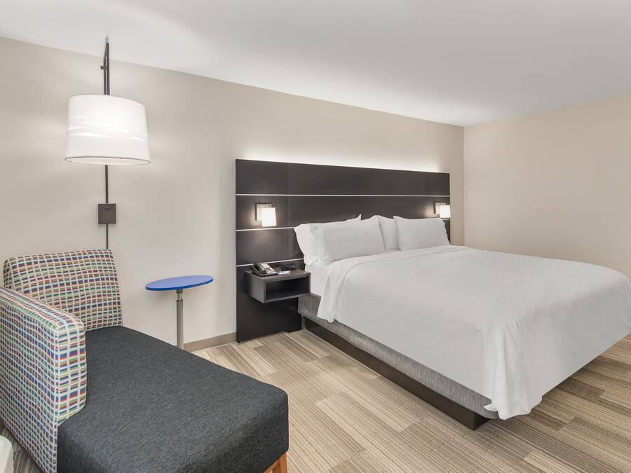 A hotel room with a bed, a chair, a small table, and a wall-mounted lamp.
