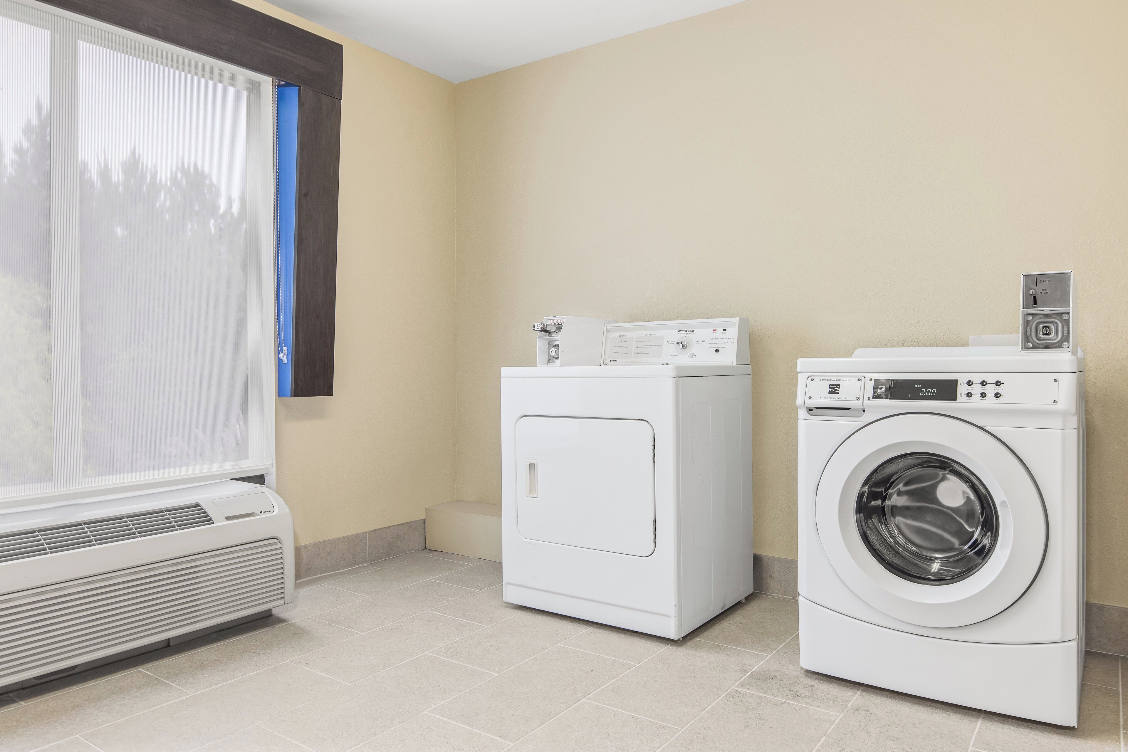 A laundry room with a front-loading washer and dryer, a vented air conditioner, and a window with blinds.