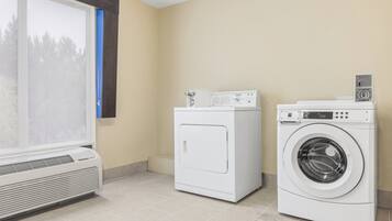 A laundry room with a front-loading washer and dryer, a vented air conditioner, and a window with blinds.