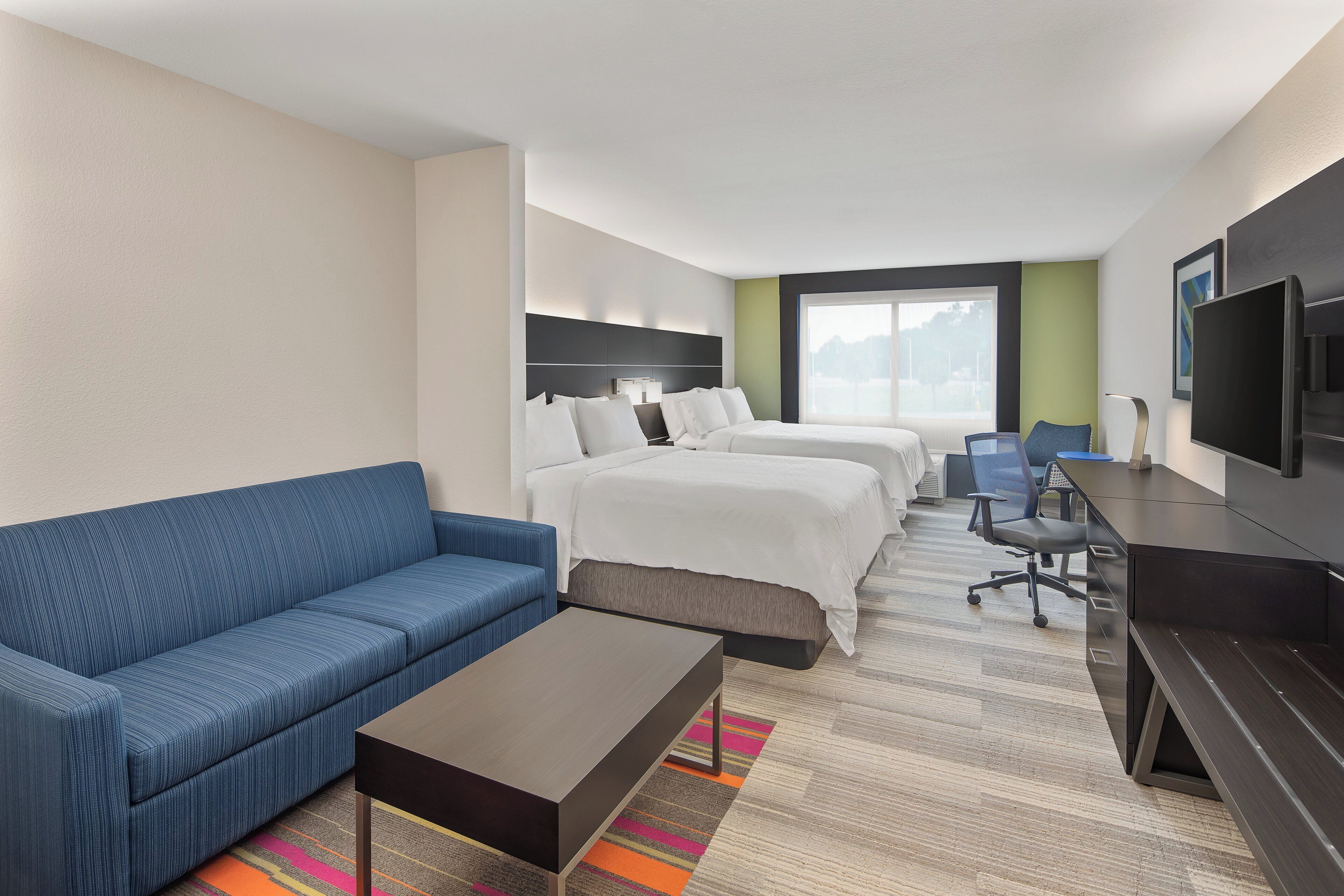 A hotel room with a sofa, two beds, a desk, a TV, and a large window.