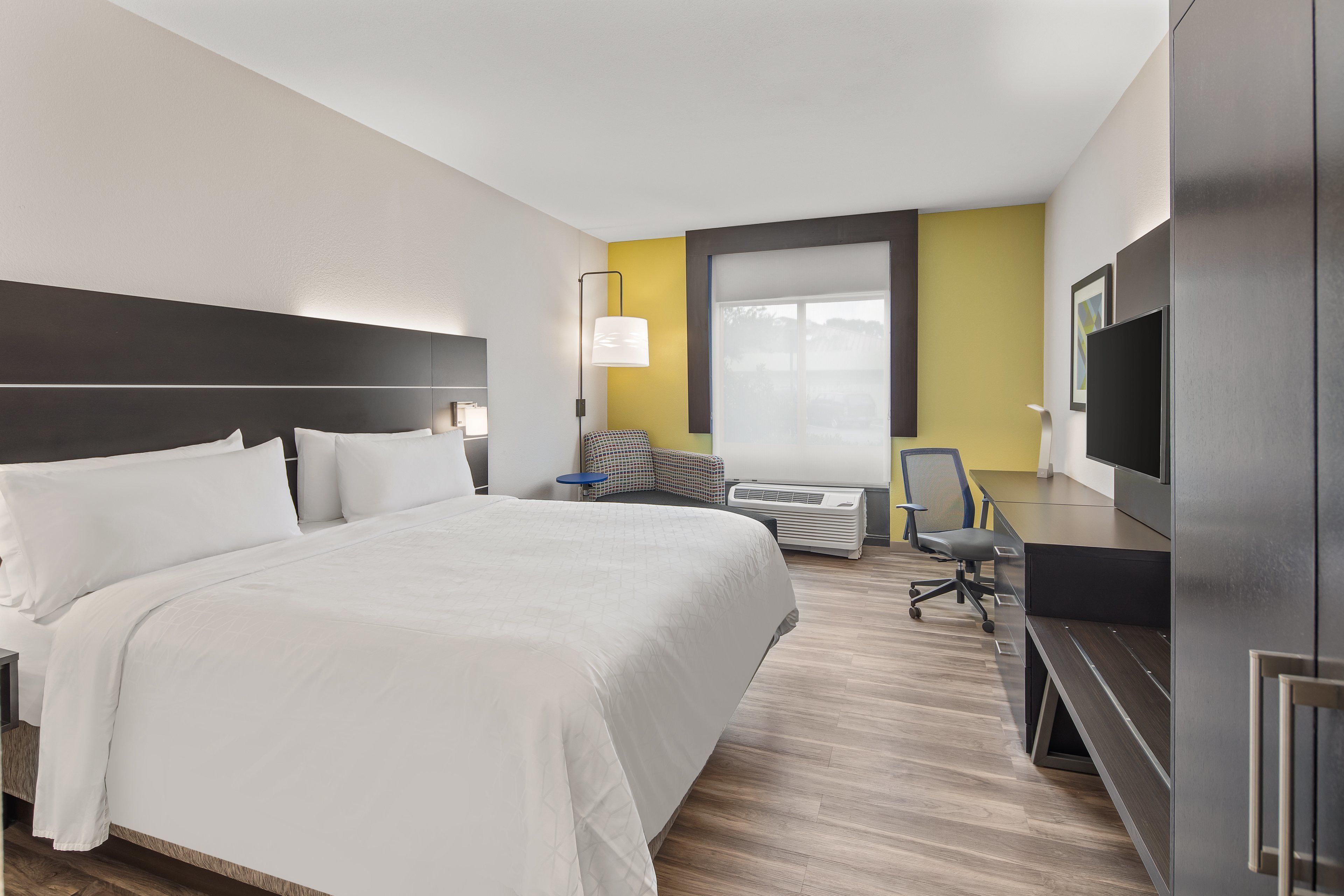 A hotel room with a large bed, a desk, a chair, a TV, and an air conditioning unit.