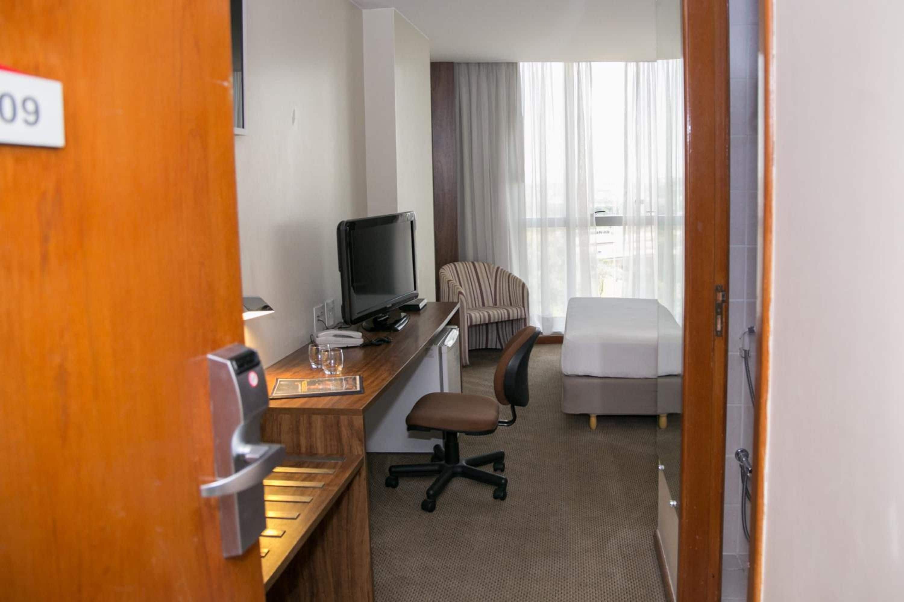 Et hotelværelse med skrivebord, computer, stol, seng og udsigt til det fri.