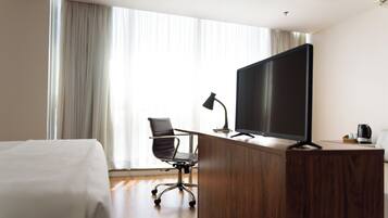 A hotel room with a wooden desk, a large flat-screen TV, a black office chair, a lamp, and a bed with a white comforter.