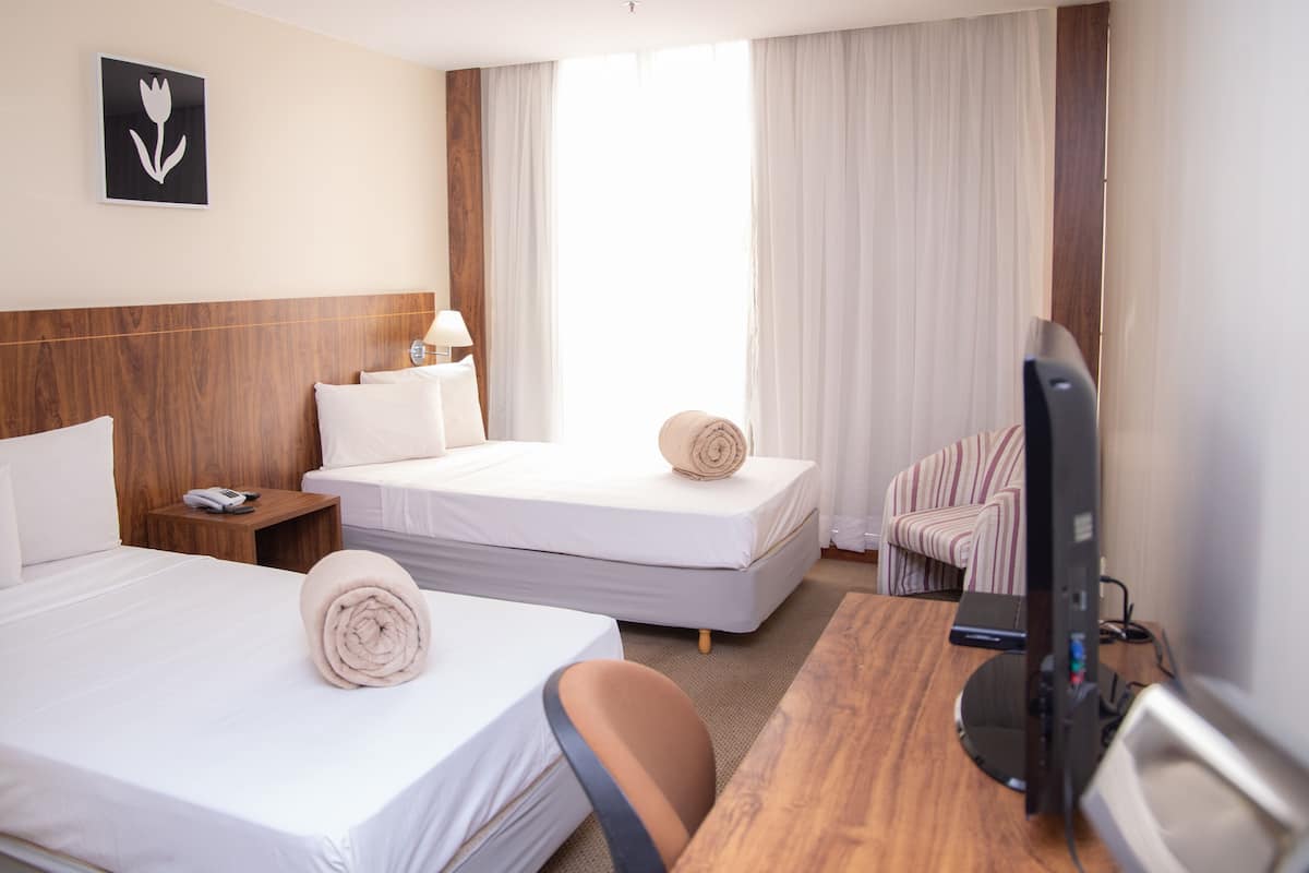 Habitación de hotel con dos camas, un escritorio con computadora, una silla, un televisor y una ventana con cortinas.