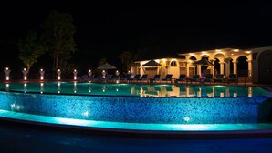 Una piscina nocturna con sillones de descanso y un edificio con arcos al fondo.